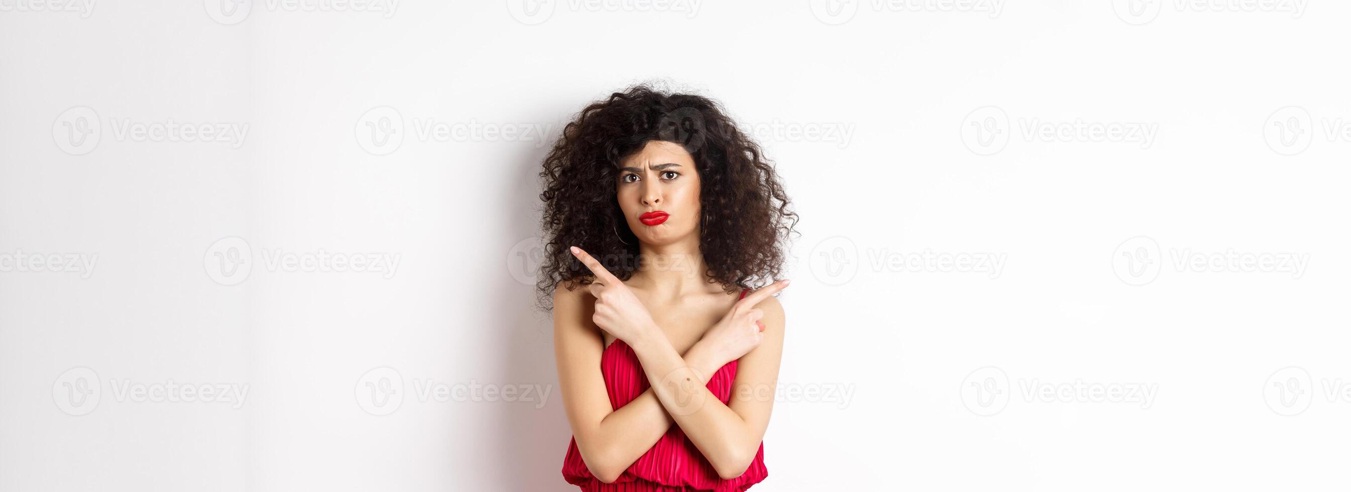 Doubtful and sad young woman in red dress, pointing fingers sideways and sulking, cant decide between products, need help with choice, standing over white background photo