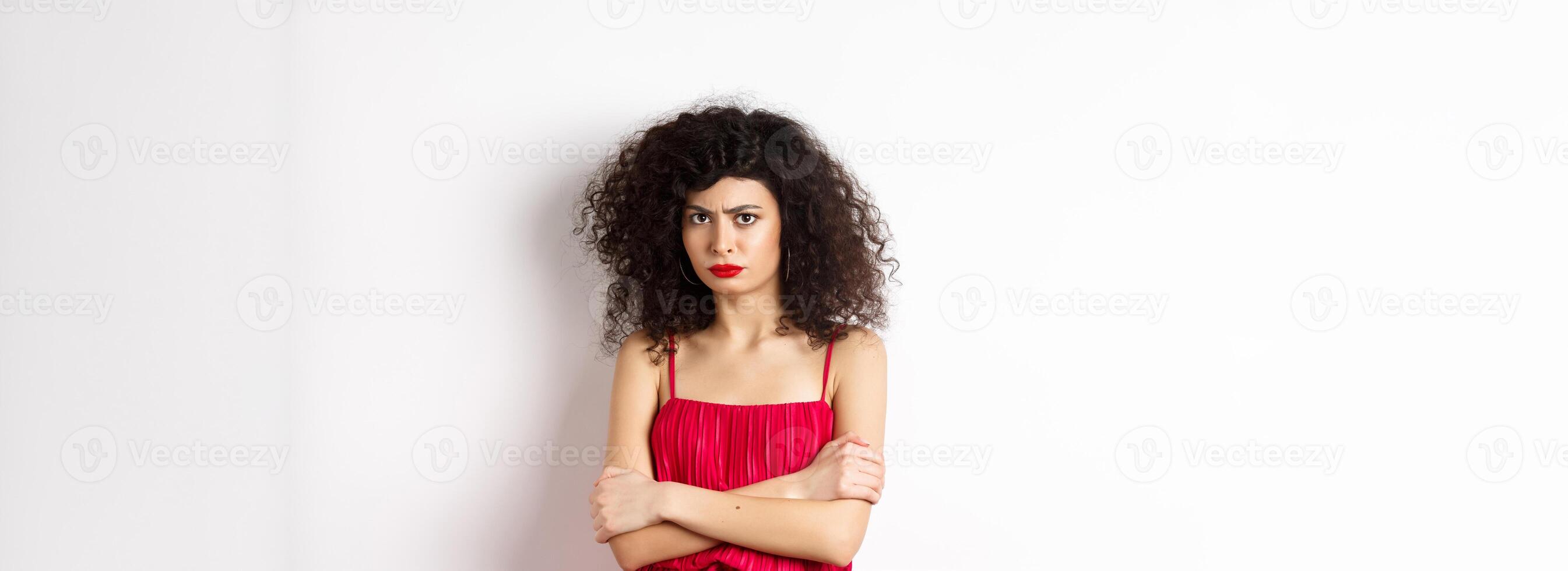 Sad and offended caucasian woman in red dress frowning, cross arms on chest and sulking, feeling mad at you, standing over white background photo