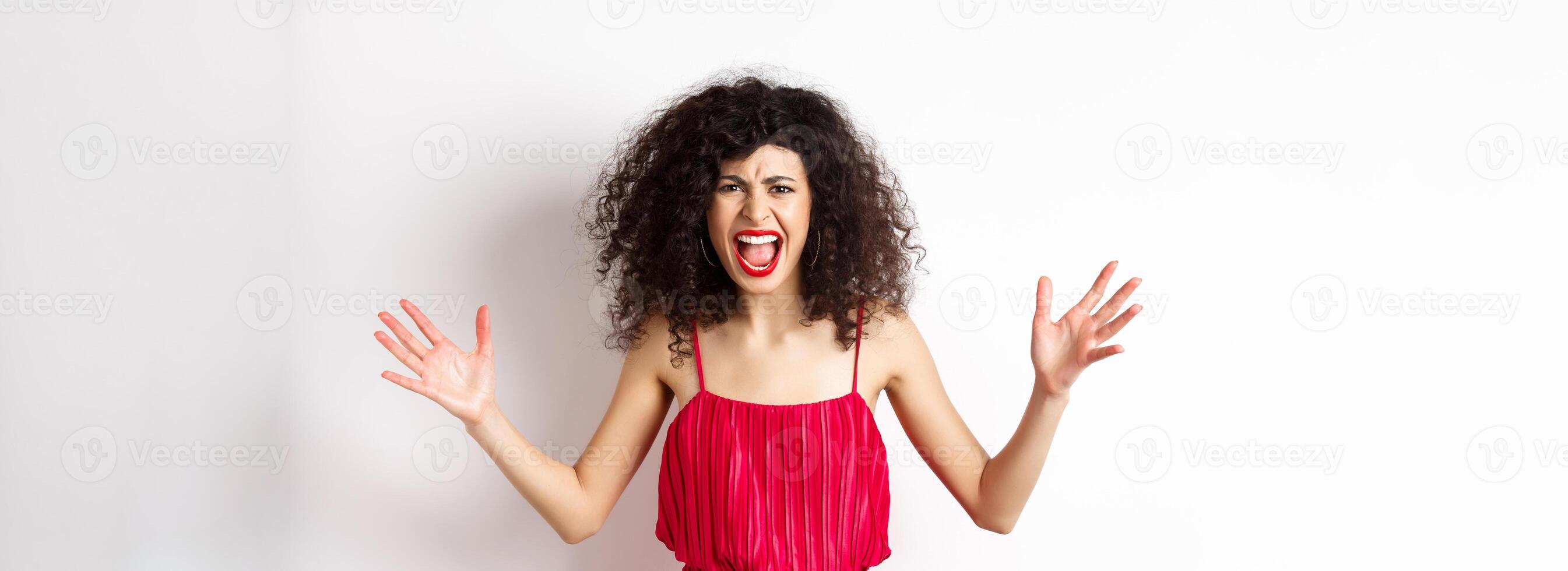 Angry young woman with curly hair, wearing red dress, screaming and having an argument, looking with hatred and anger, standing over white background photo