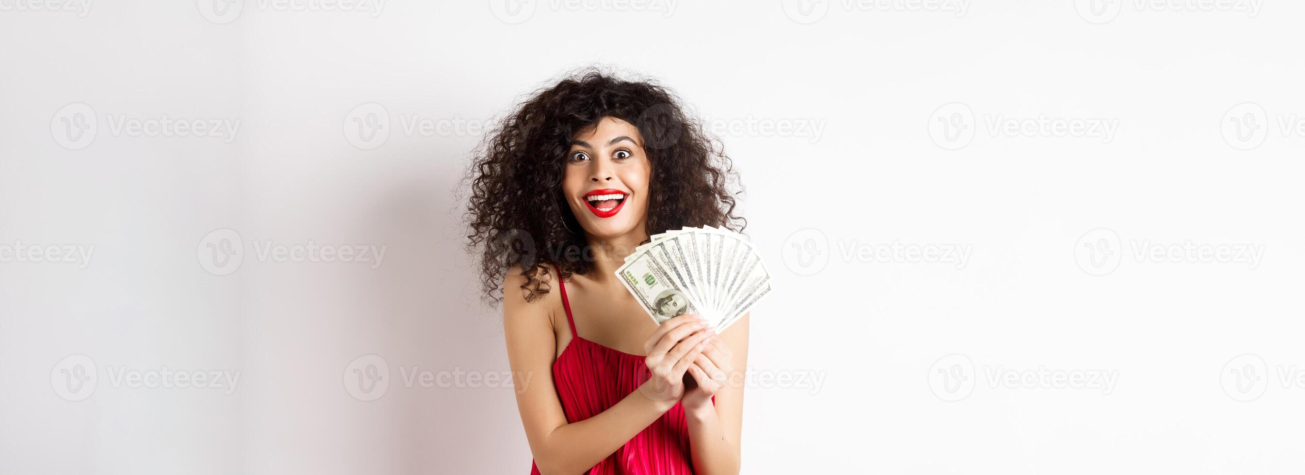 Excited woman in red dress winning money, showing dollar bills and smiling happy, standing on white background photo