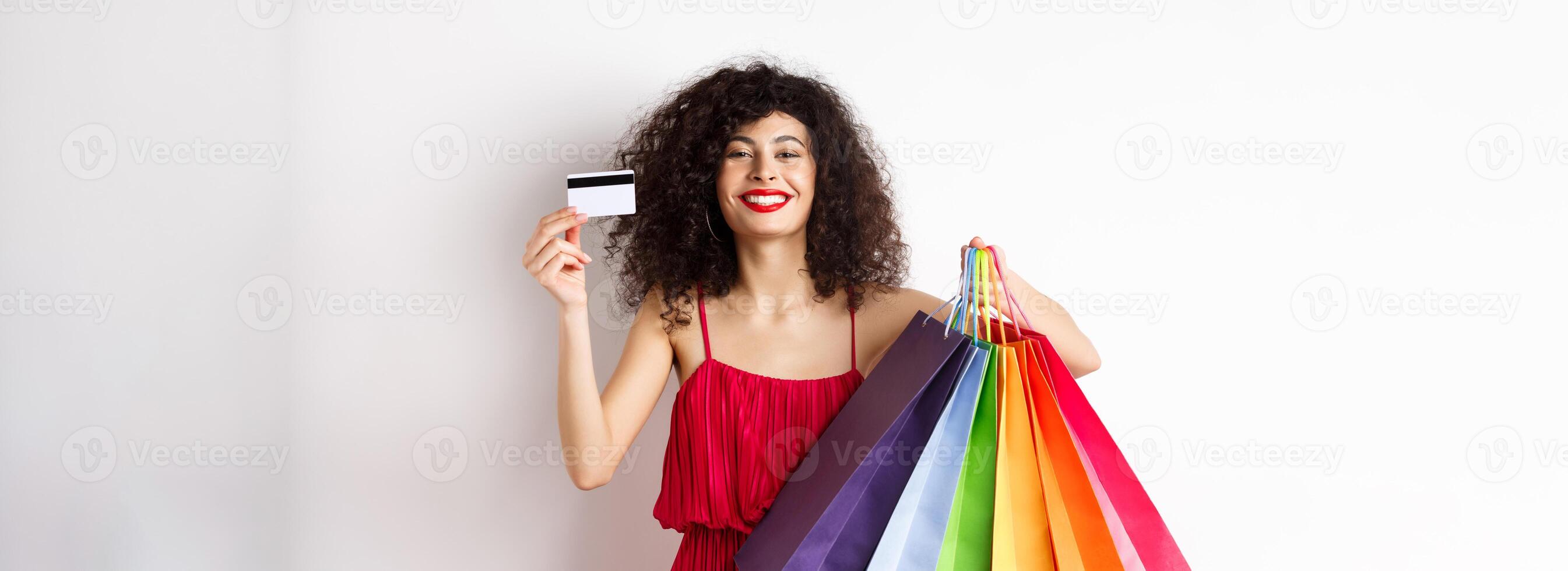 Happy elegant woman in red dress, showing shopping bags and plastic credit card, smiling pleased, standing on white background photo