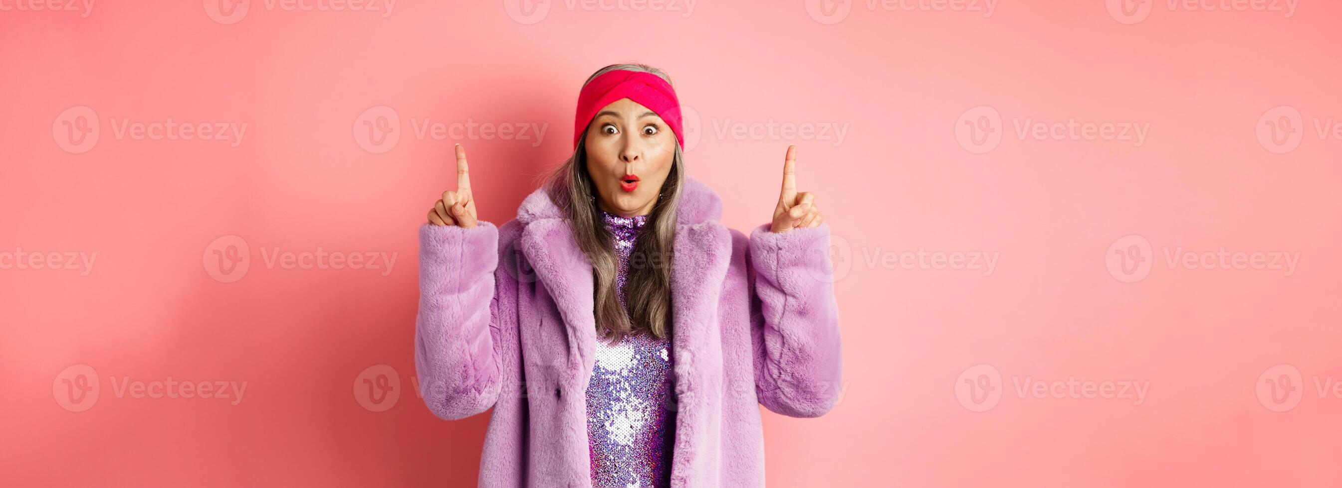 Wow check this out. Amazed asian senior woman pointing fingers up, staring at camera impressed, showing fashion or beauty advertisement, pink background photo