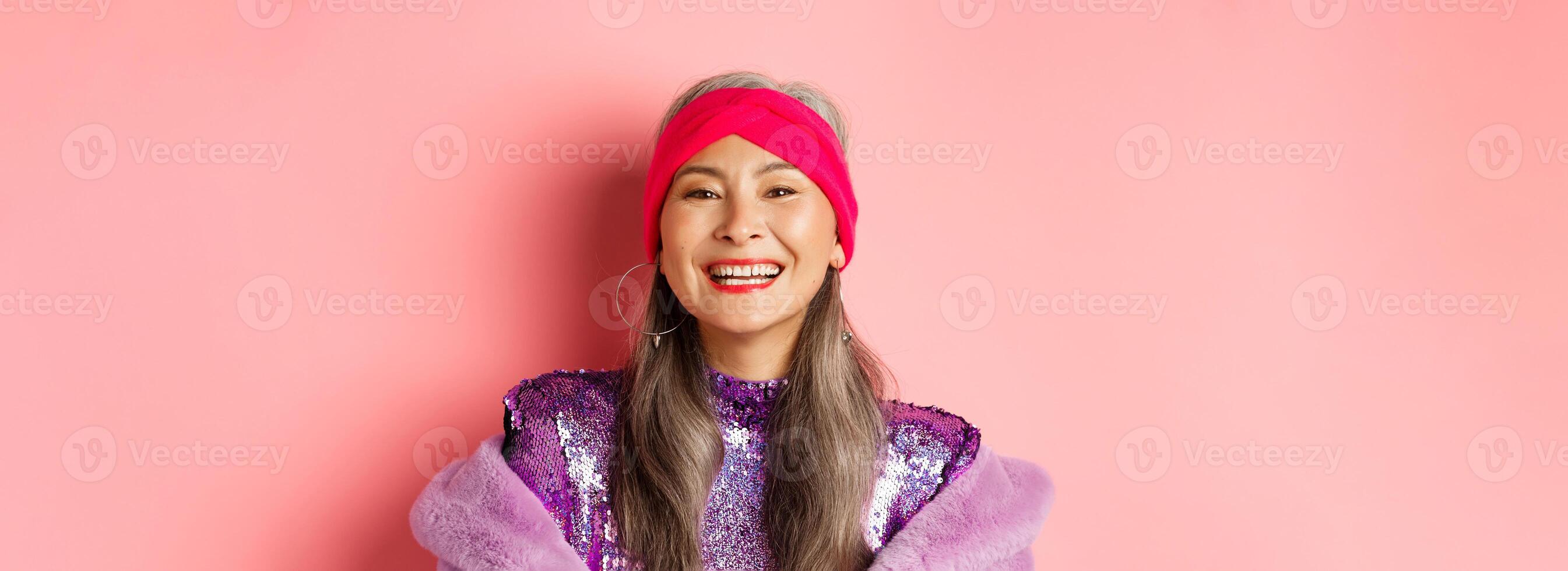 Close-up of happy asian senior lady in trendy purple outfit, laughing and smiling at camera, having fun on pink background photo