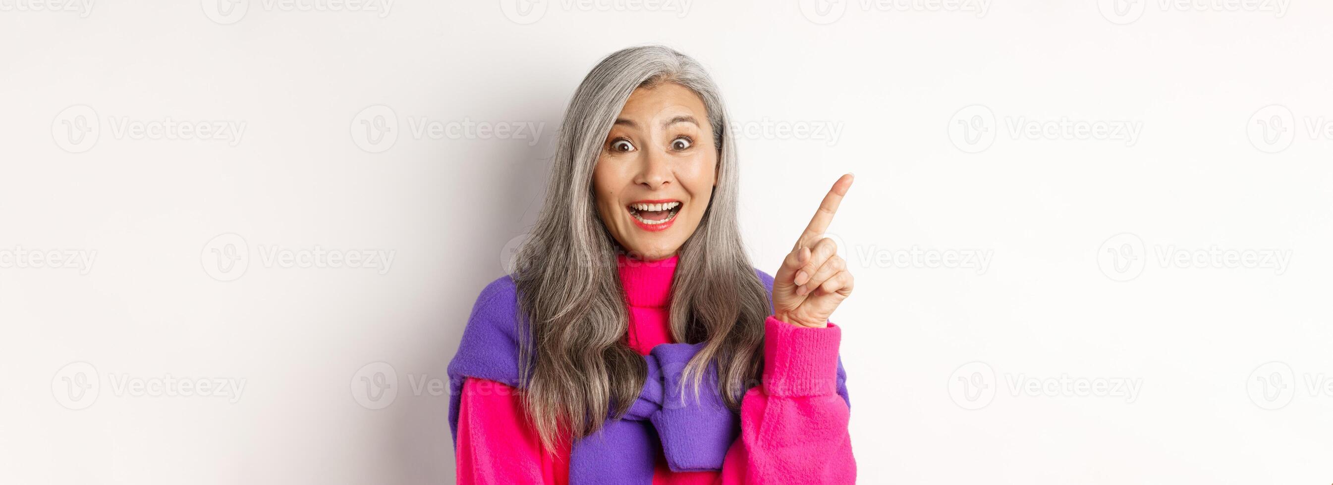 Close up of beautiful asian female model pointing finger at upper left corner, smiling and looking happy at camera, showing promo deal, white background photo