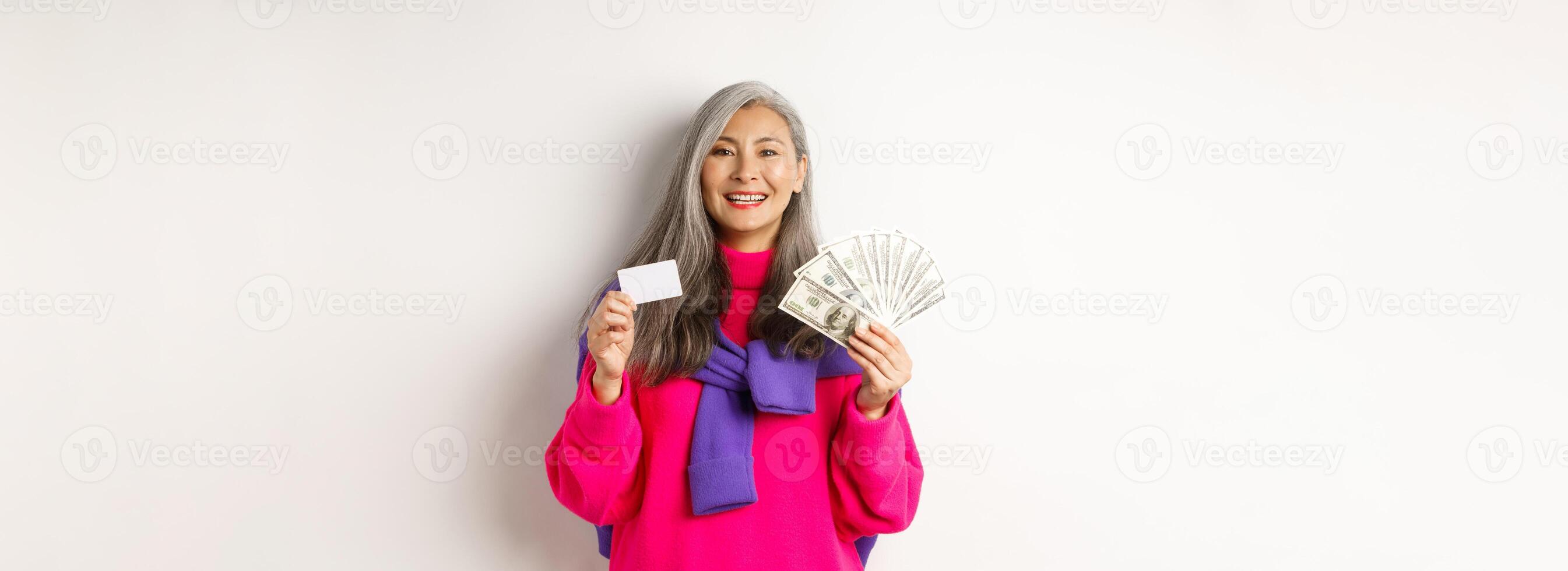 Shopping concept. Stylish asian senior woman with grey hair, showing money dollars and plastic credit card, standing over white background photo
