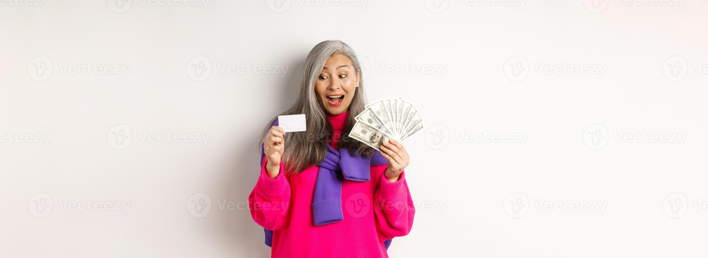 Shopping concept. Lucky asian senior woman looking amazed at credit card and showing money dollars, standing over white background photo