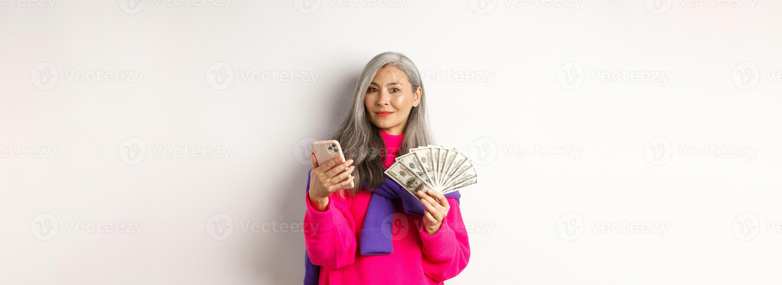Modern asian senior lady holding money and smartphone, order something online, standing over white background photo