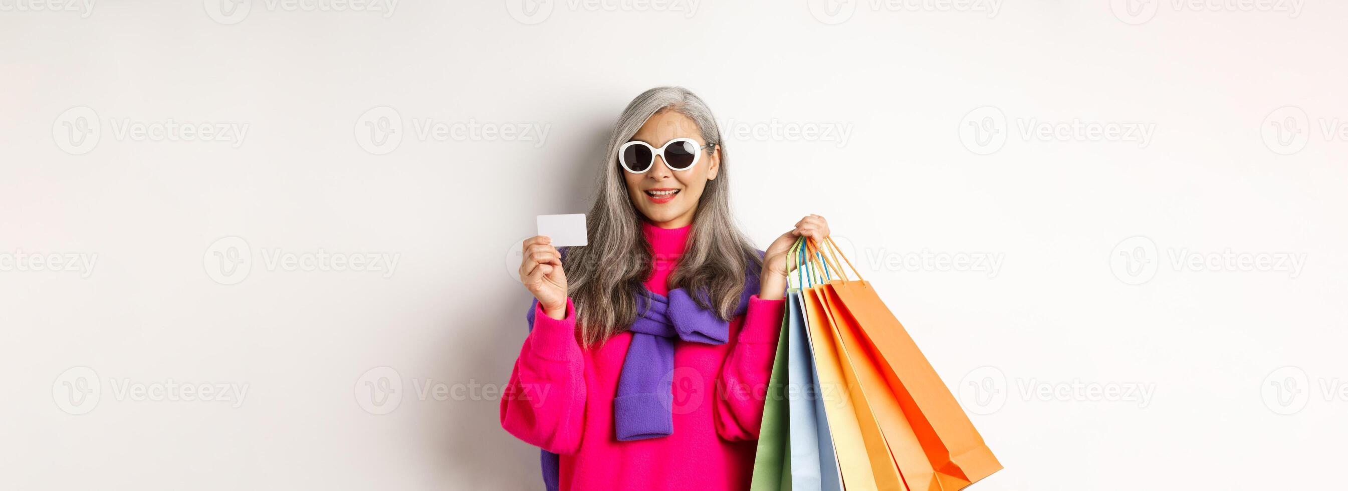Stylish senior asian woman in sunglasses going shopping on holiday sale, holding paper bags and plastic credit card, standing over white background photo