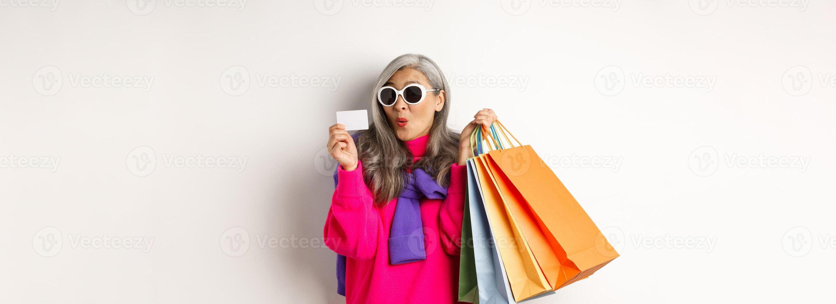 Fashionable asian old lady going shopping, holding paper bags and plastic credit card, wearing stylish sunglasses, standing over white background photo