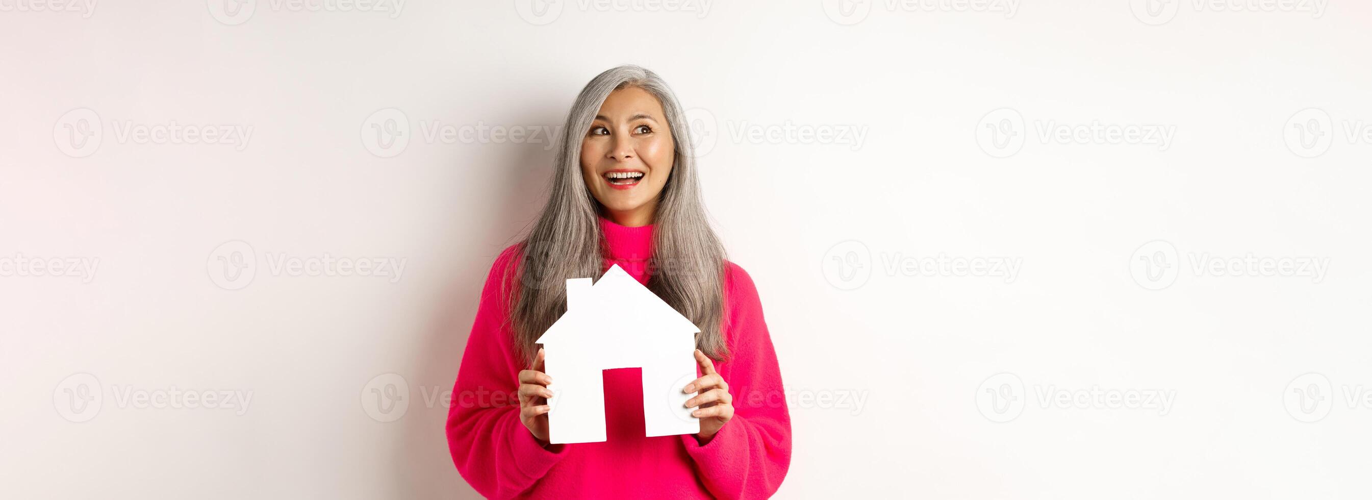 Real estate. Beautiful asian adult woman with grey hair, dreaming about buying property, showing paper house model and looking at upper left corner, white background photo