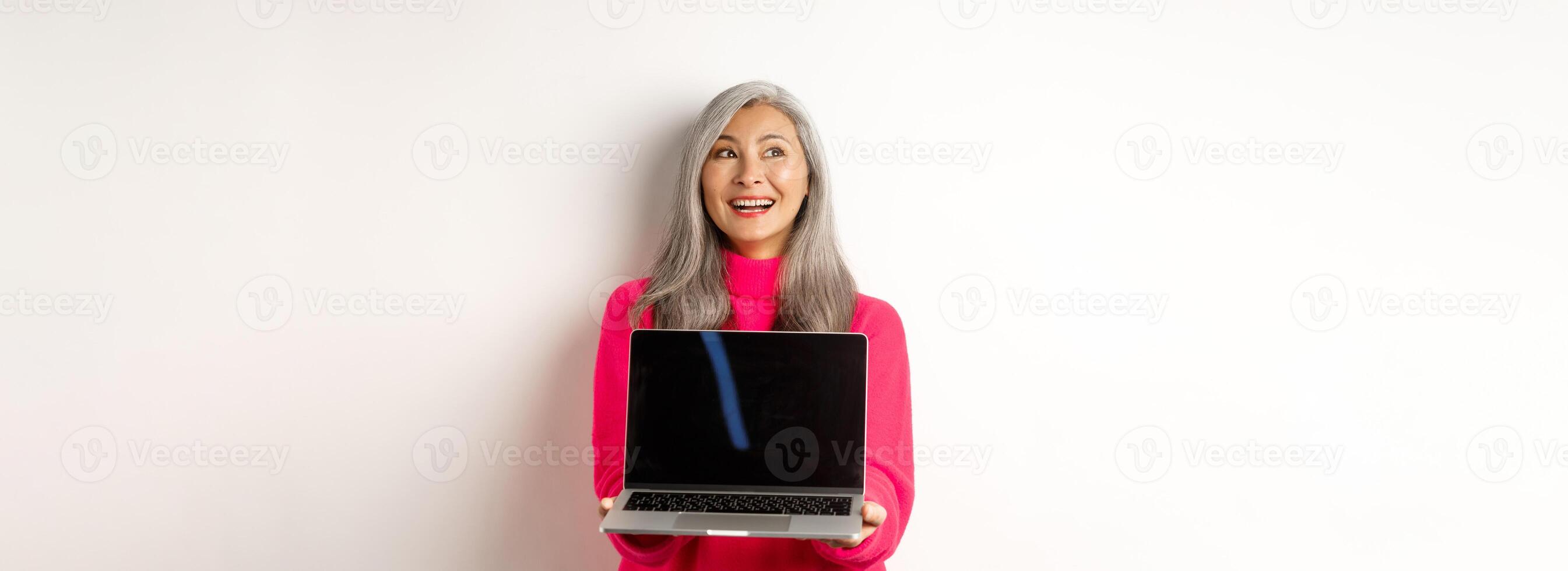 E-commerce concept. Happy beautiful asian senior woman with grey hair, looking aside and smiling, showing laptop black screen, standing over white background photo
