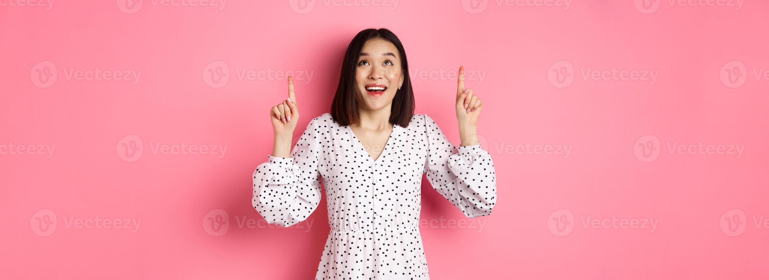 Amazed brunette girl in dress looking, pointing fingers up, showing logo with wondered smile, standing over pink background photo