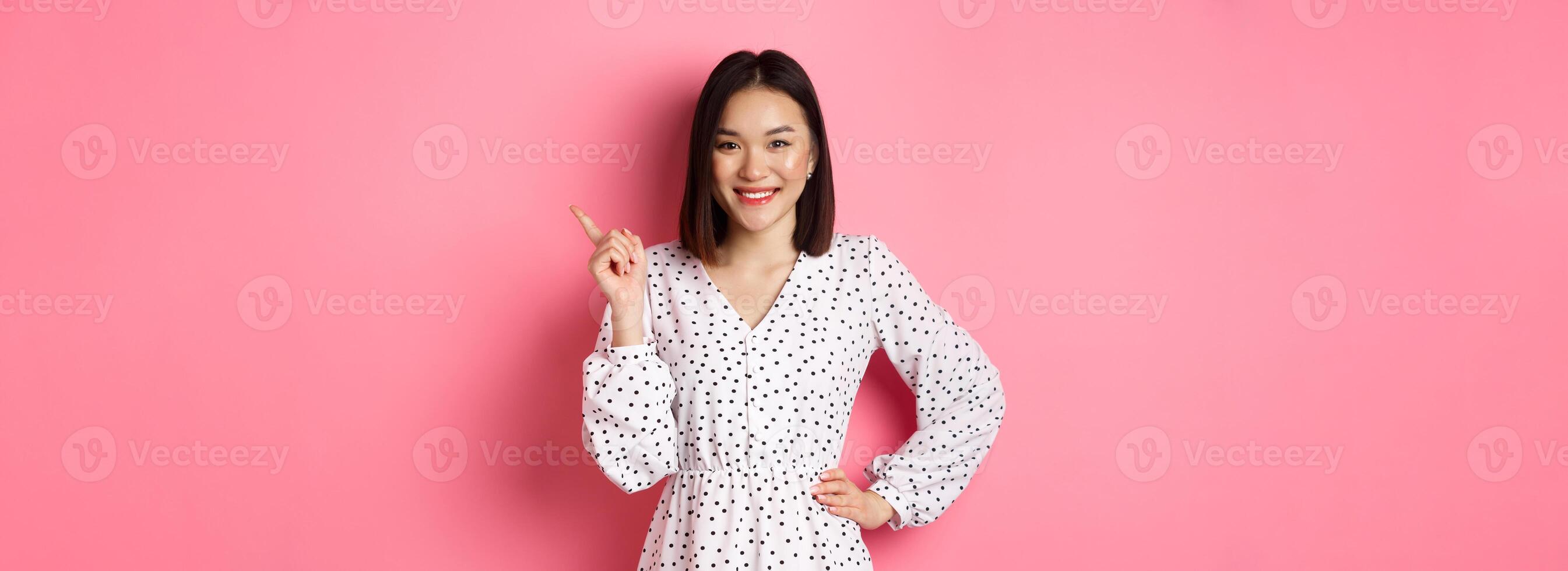 Beautiful asian female model smiling, pointing finger at upper right corner copy space, showing advertisement banner, standing over pink background photo