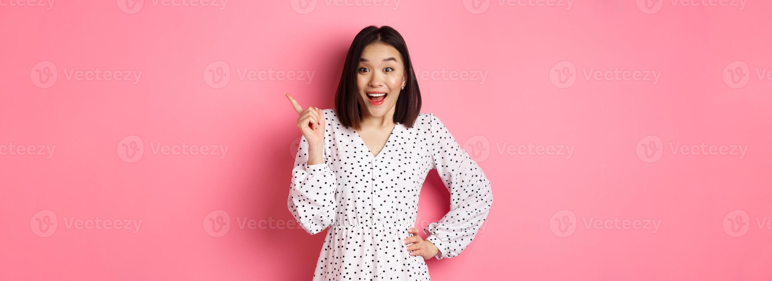 Excited pretty asian girl pointing at upper right corner, gasping amazed and looking at camera, discuss store promo offer, standing over pink background photo