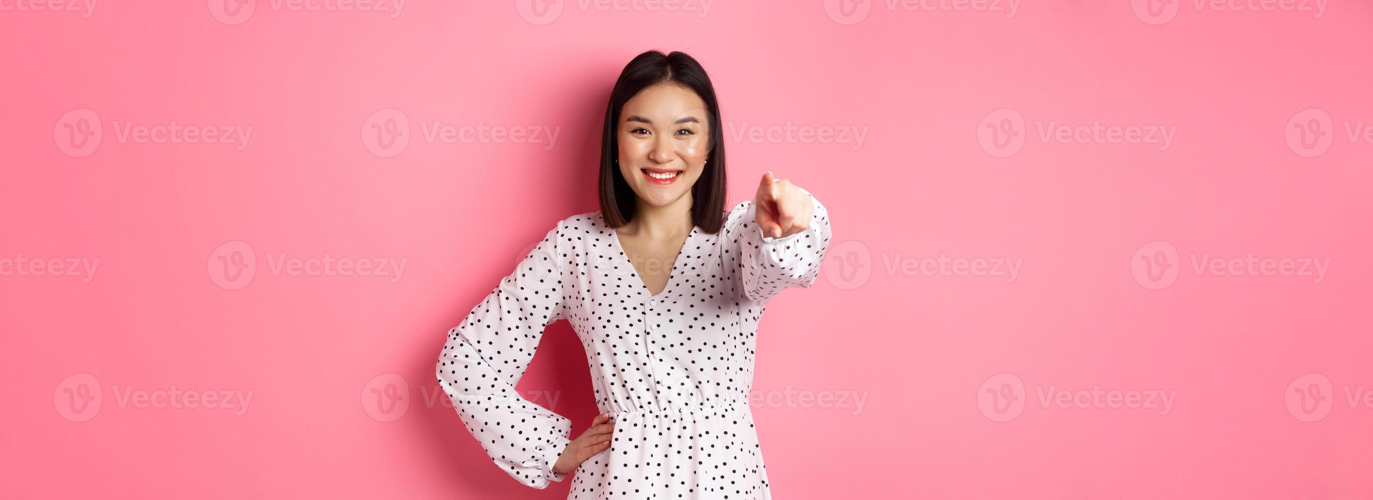 Confident asian woman in dress pointing at you, smiling and staring at camera, standing against pink background photo