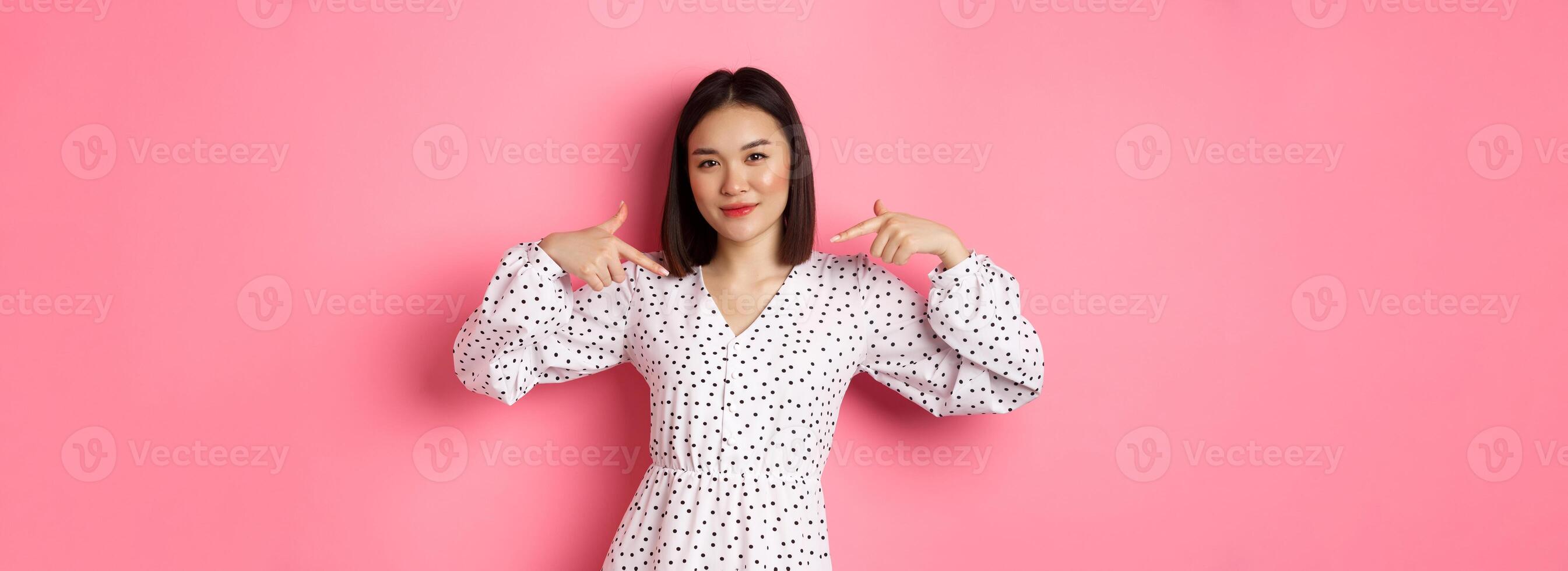 Beautiful asian woman looking confident, pointing at herself and smiling, showing logo on chest, standing over pink background photo