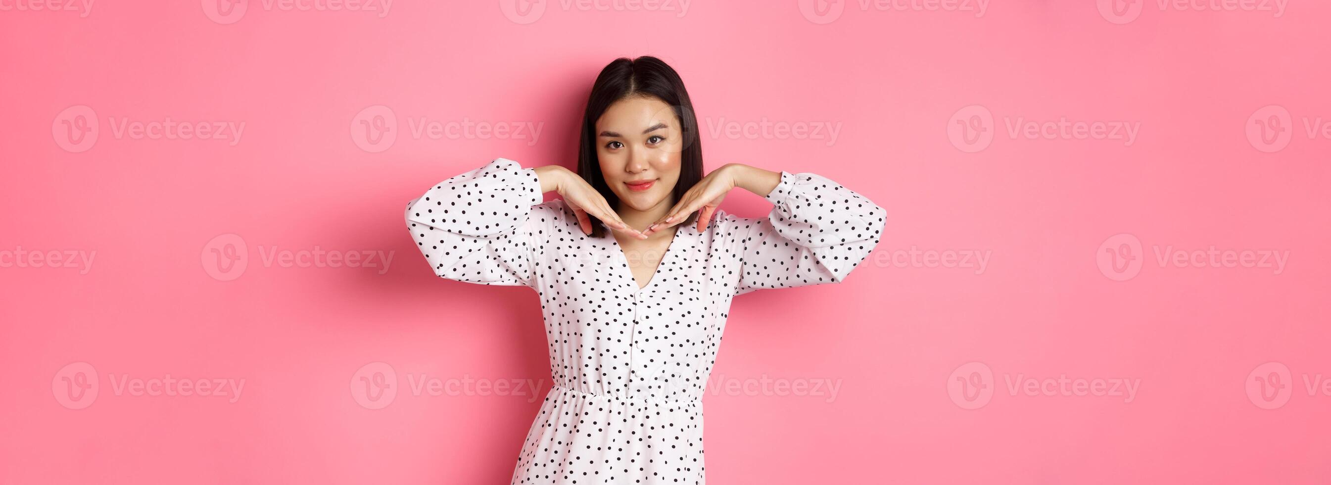 Beautiful asian woman in trendy dress making cute face, holding hands near jawline and gazing coquettish at camera, standing over pink background photo