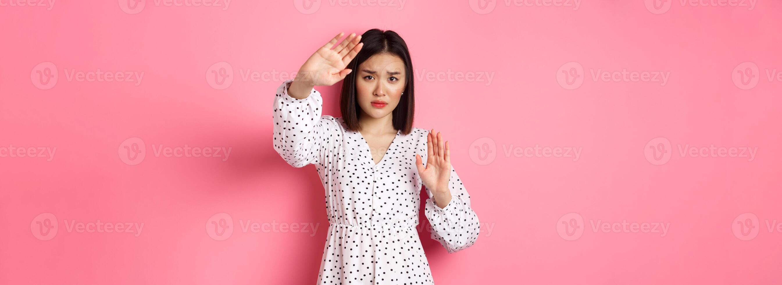 Timid and scared asian woman defending herself, raising arms in protection, victim being attacked, standing over pink background photo