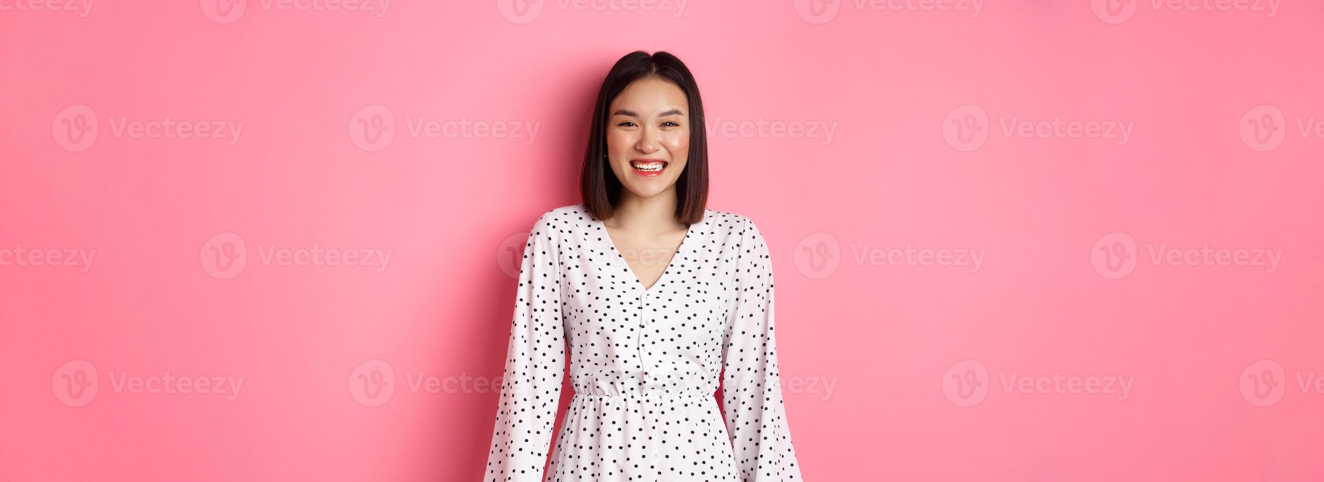 Happy korean woman in dress looking at camera, smiling and laughing with sincere expression, standing over pink background photo