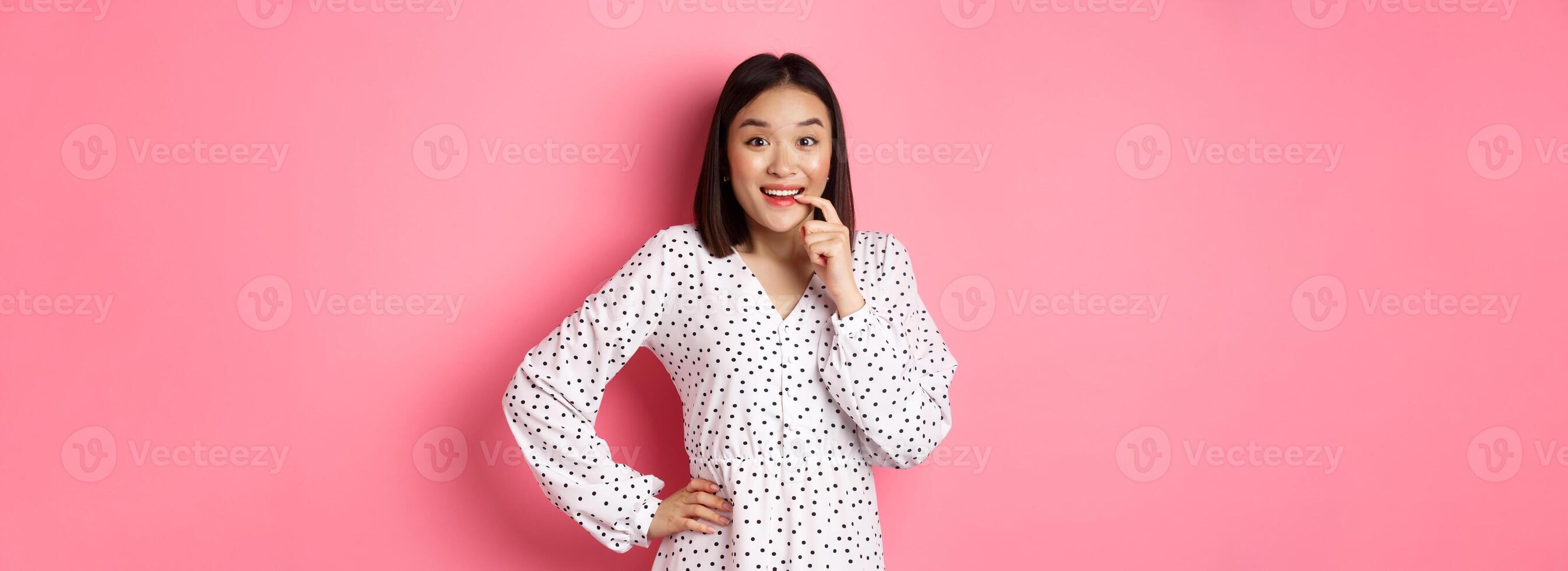 Image of beautiful young lady in dress, biting finger and smiling, staring with temptation and amazement, want buy something, standing over pink background photo