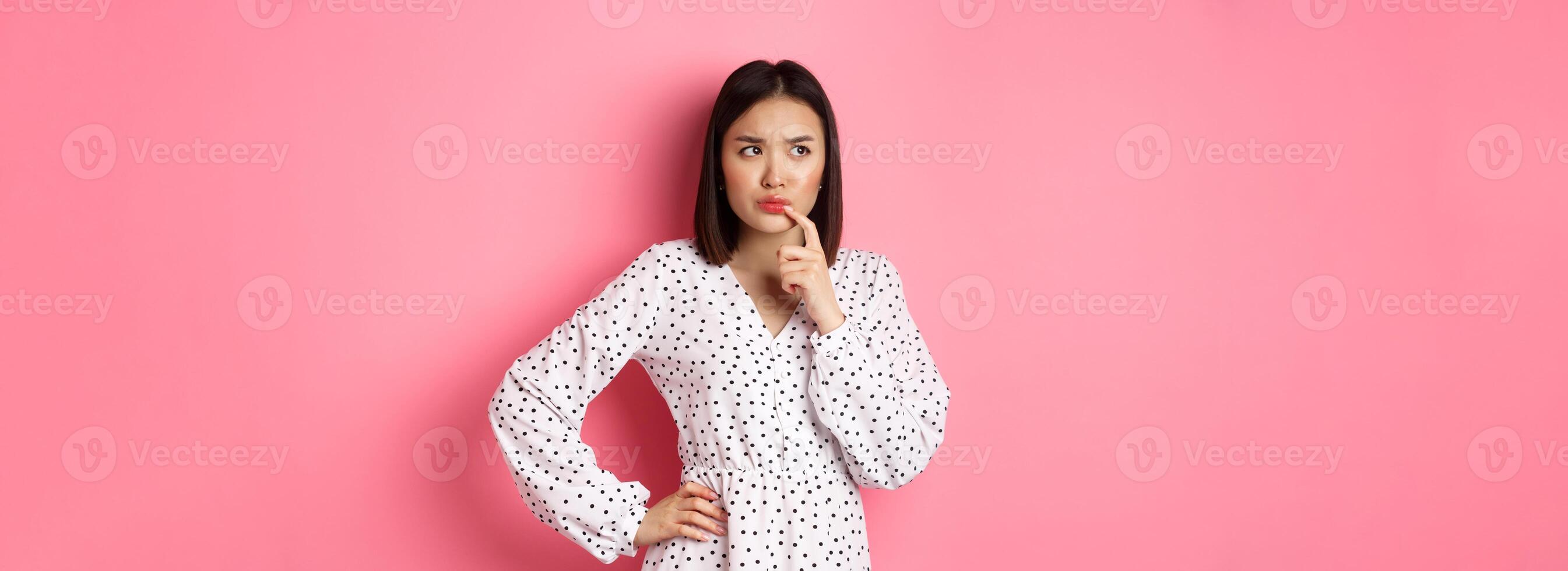 Troubled asian girl making decision, frowning and touching lip while thinking, looking unsure at upper left corner and choosing, standing over pink background photo