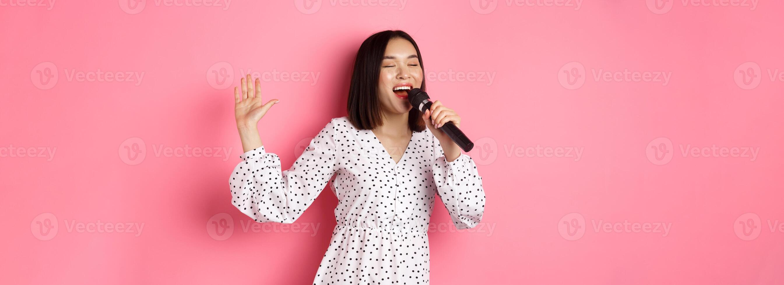 Happy asian woman dancing and singing in microphone, performing at karaoke, standing over pink background photo
