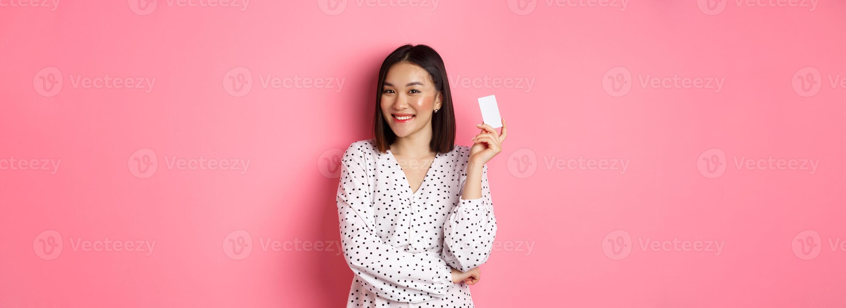 Shopping concept. Confident and happy asian woman holding credit card and smiling satisfied, standing against pink background photo