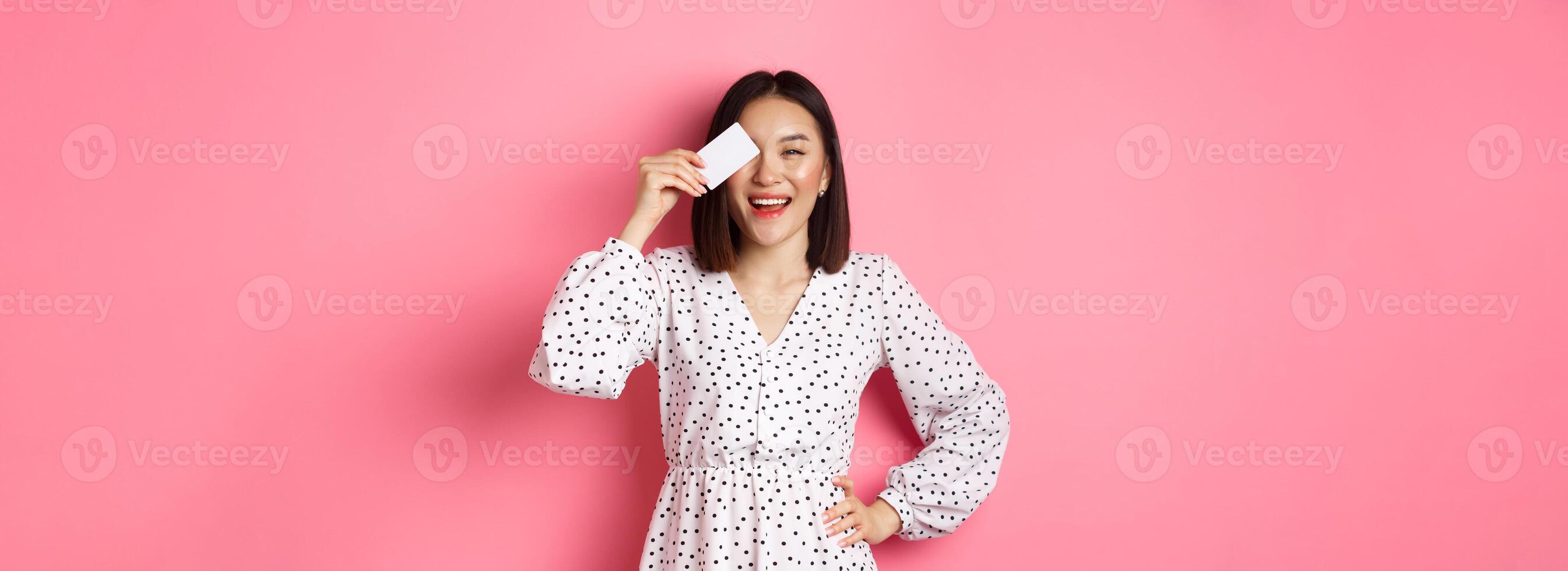 Shopping concept. Pretty asian woman holding bank credit card and smiling, standing over pink background photo