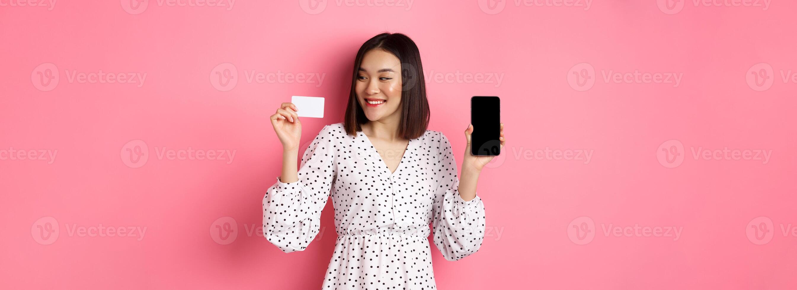 Happy young asian woman internet shopping, showing smartphone screen and looking satisfied at credit card, standing over pink background photo