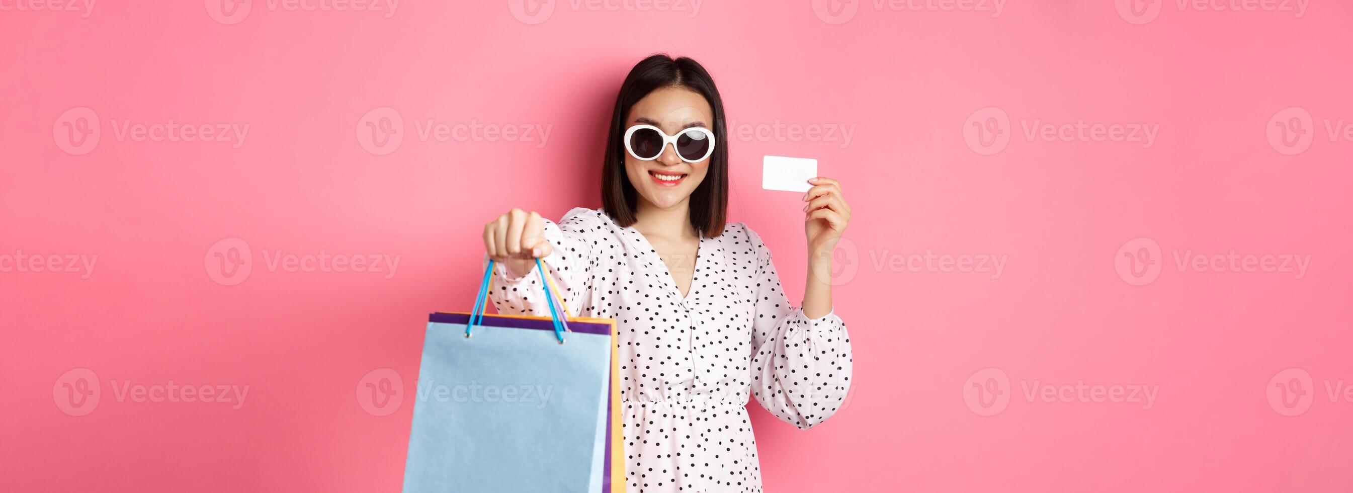 Beautiful asian woman in sunglasses going shopping, holding bags and showing credit card, standing over pink background photo