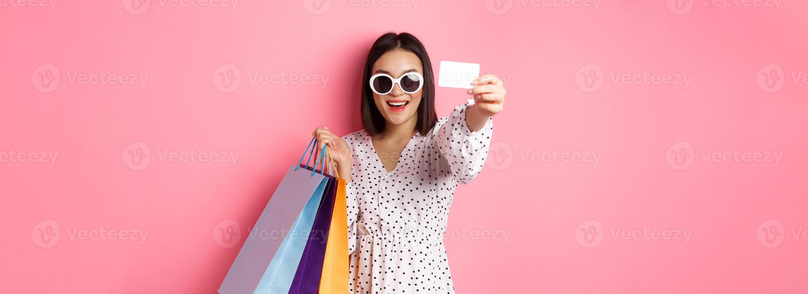 Beautiful asian woman in sunglasses going shopping, holding bags and showing credit card, standing over pink background photo