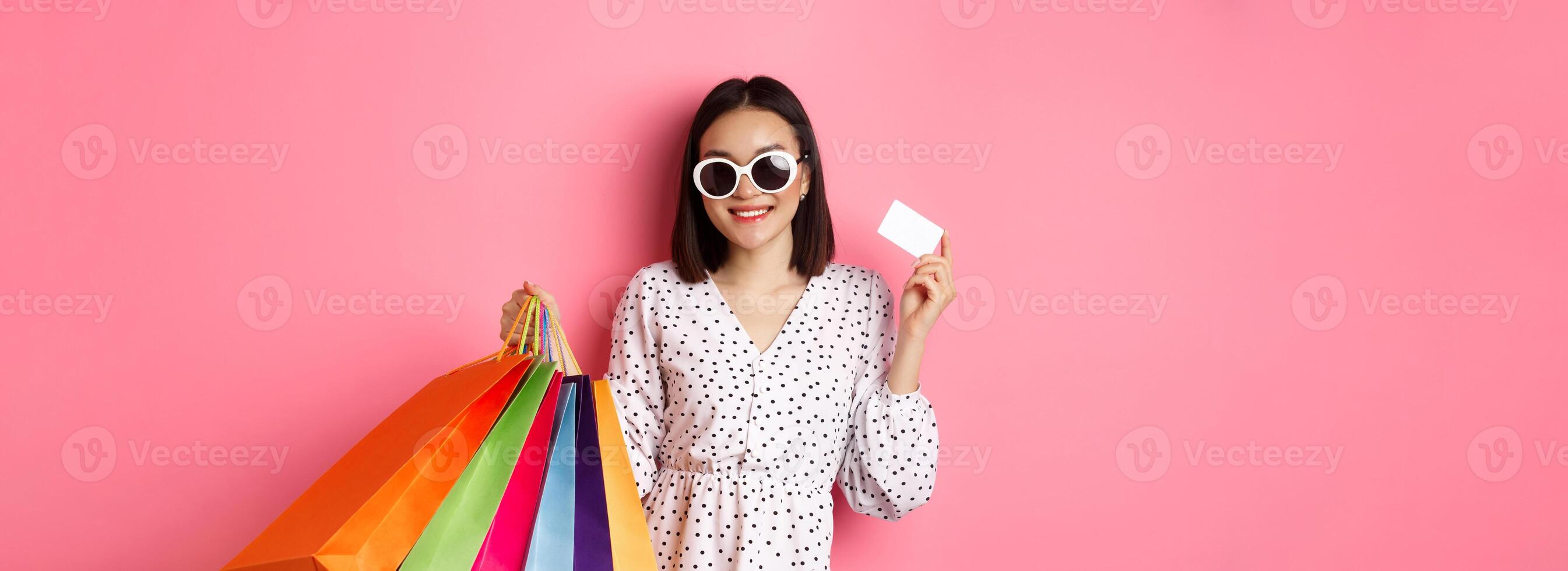 Beautiful asian woman in sunglasses going shopping, holding bags and showing credit card, standing over pink background photo