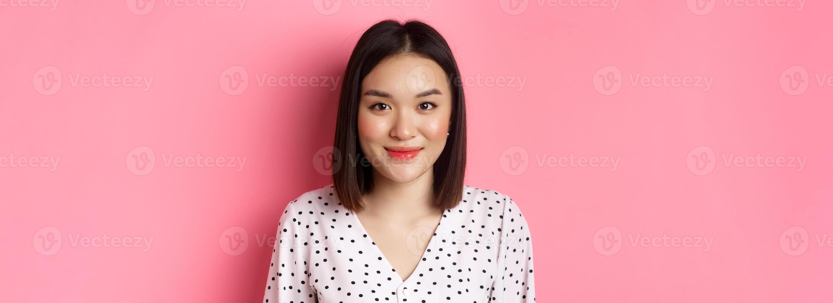Close-up of cute asian female model looking at camera, blushing and smiling kawaii, standing over pink background photo