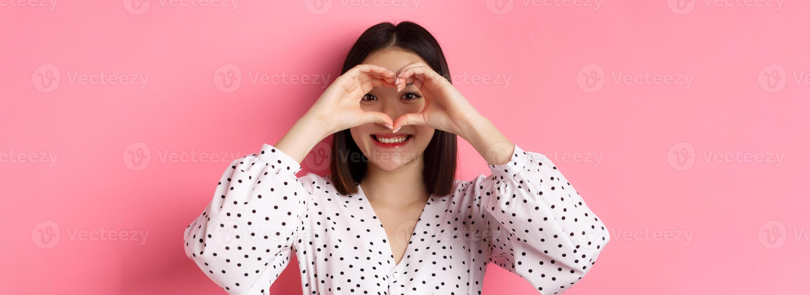Beauty and lifestyle concept. Close-up of lovely asian woman showing heart sign, smiling and feeling romantic on valentines day, standing over pink background photo