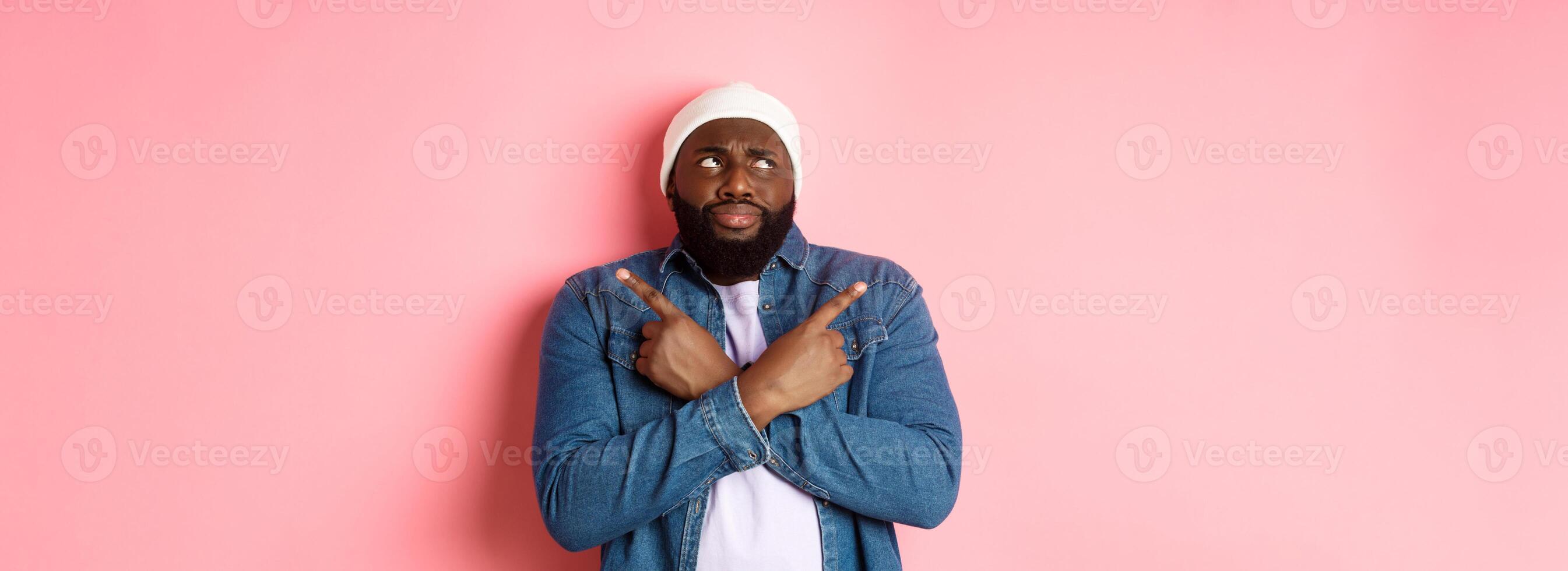 Confused Black man with beard, making choice, pointing fingers sideways and looking puzzled, standing over pink background photo
