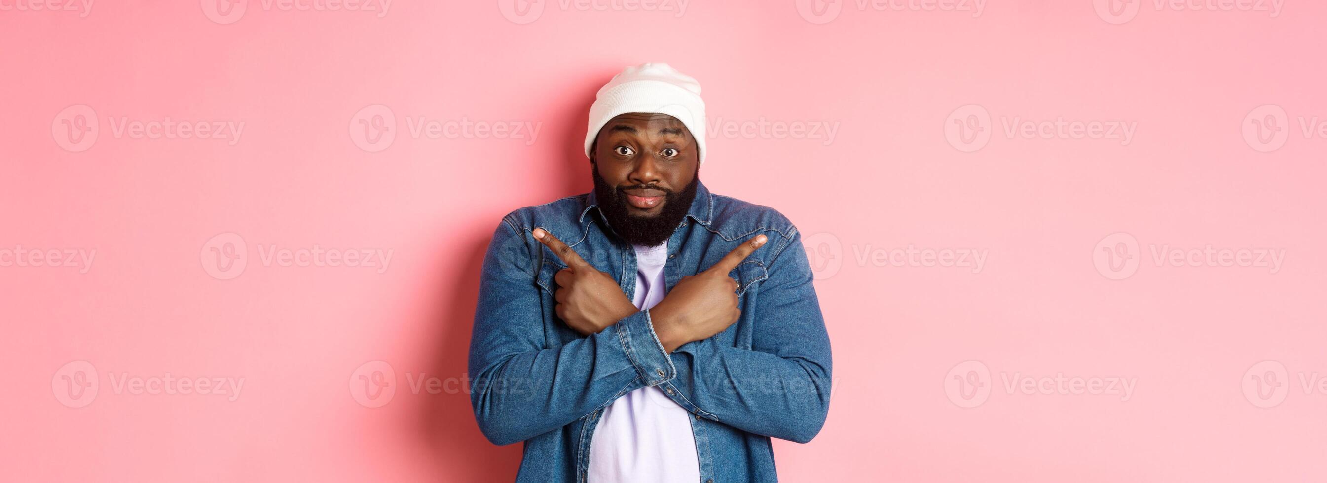 Confused Black man asking help with choice, pointing sideways and staring at camera, making decision, standing over pink background photo
