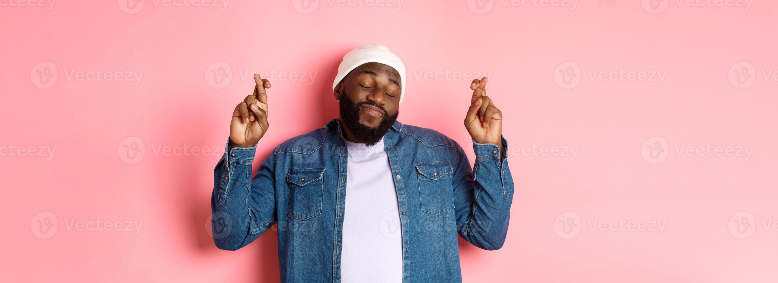 Hopeful and dreamy Black man making wish, cross fingers and smiling, standing over pink background photo
