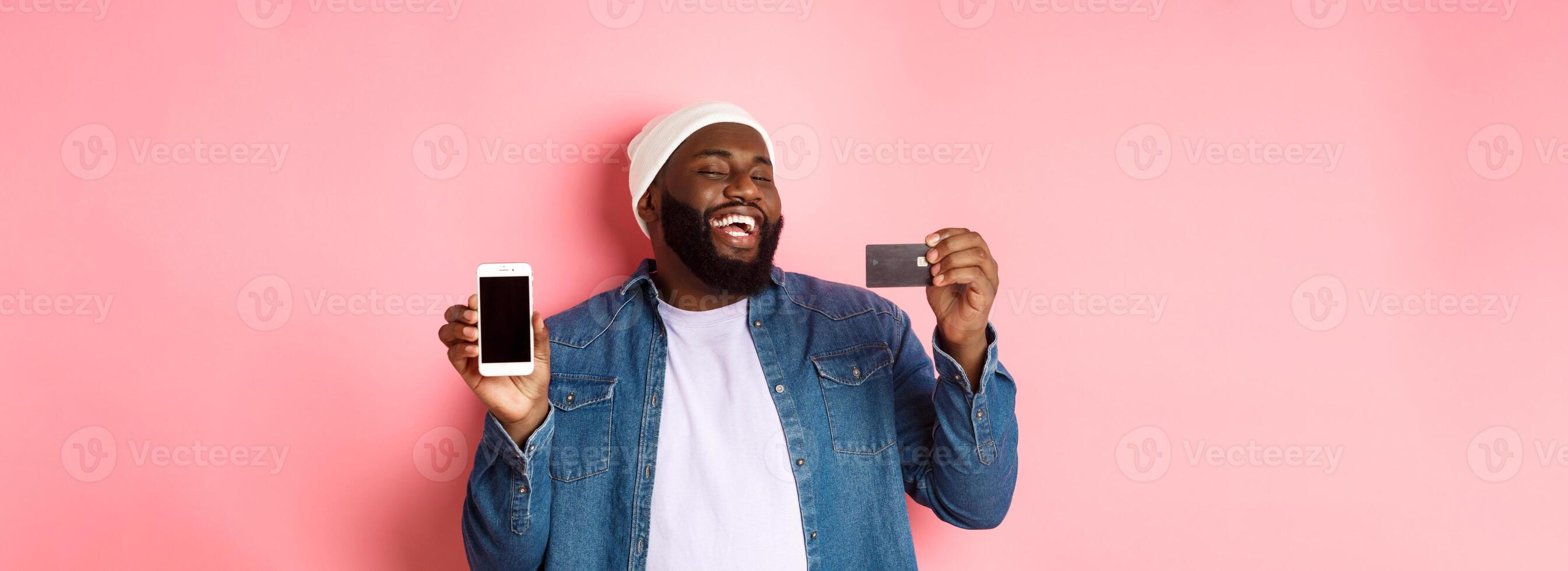 Online shopping. Happy african-american man in beanie laughing, showing credit card and mobile phone screen, standing over pink background photo