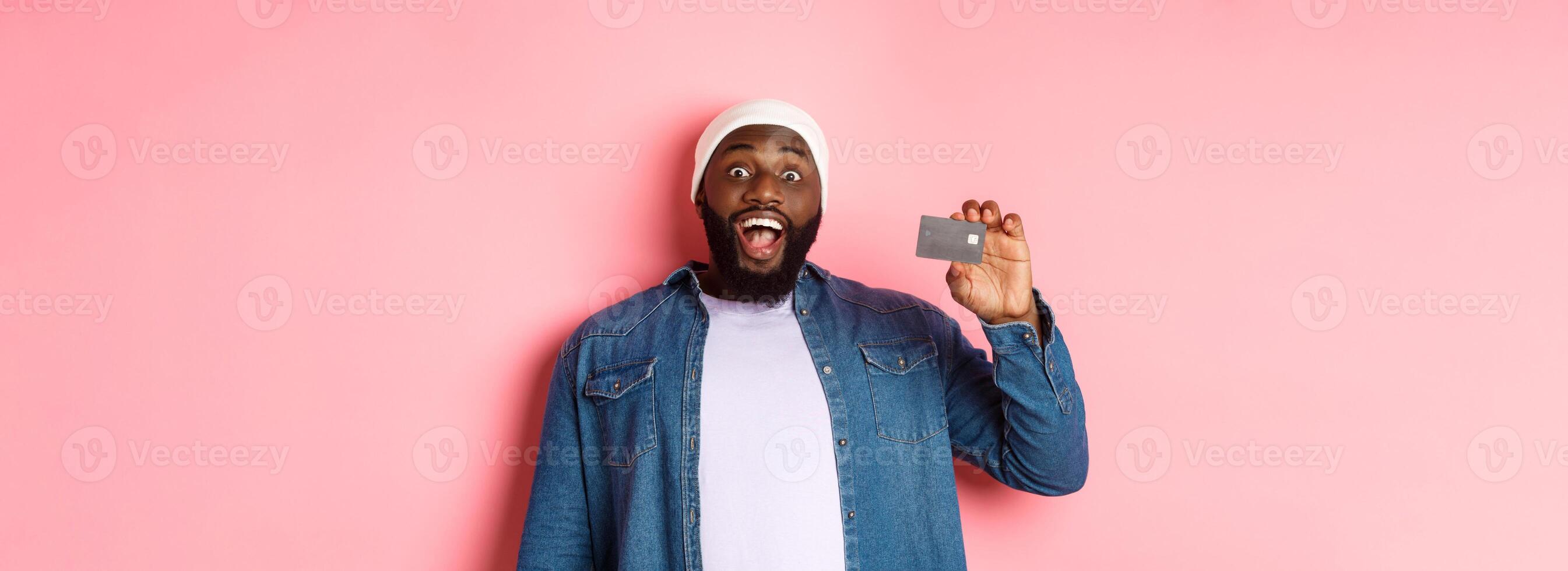 Shopping concept. Amazed Black guy showing credit card, staring at camera impressed, recommending bank, standing over pink background photo