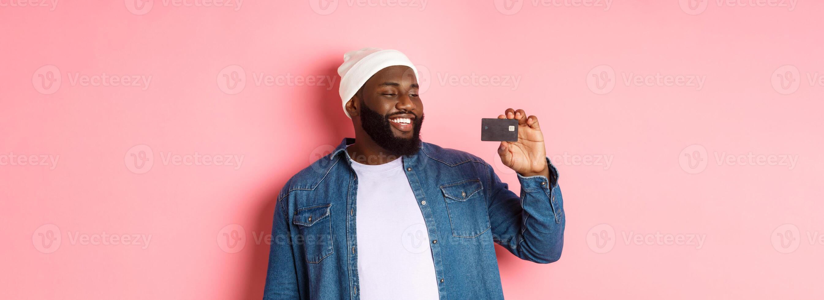 Shopping concept. Image of happy african-american man looking satisfied at credit card, recommending bank, standing over pink background photo