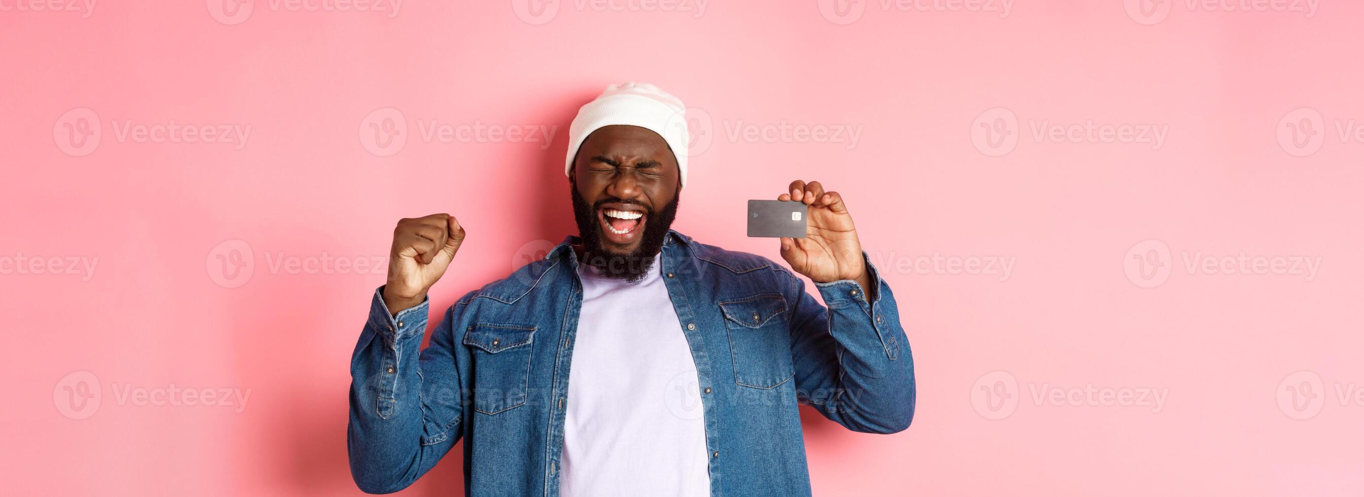 Shopping concept. Happy Black man rejoicing, scream of joy and showing credit card, standing over pink background photo