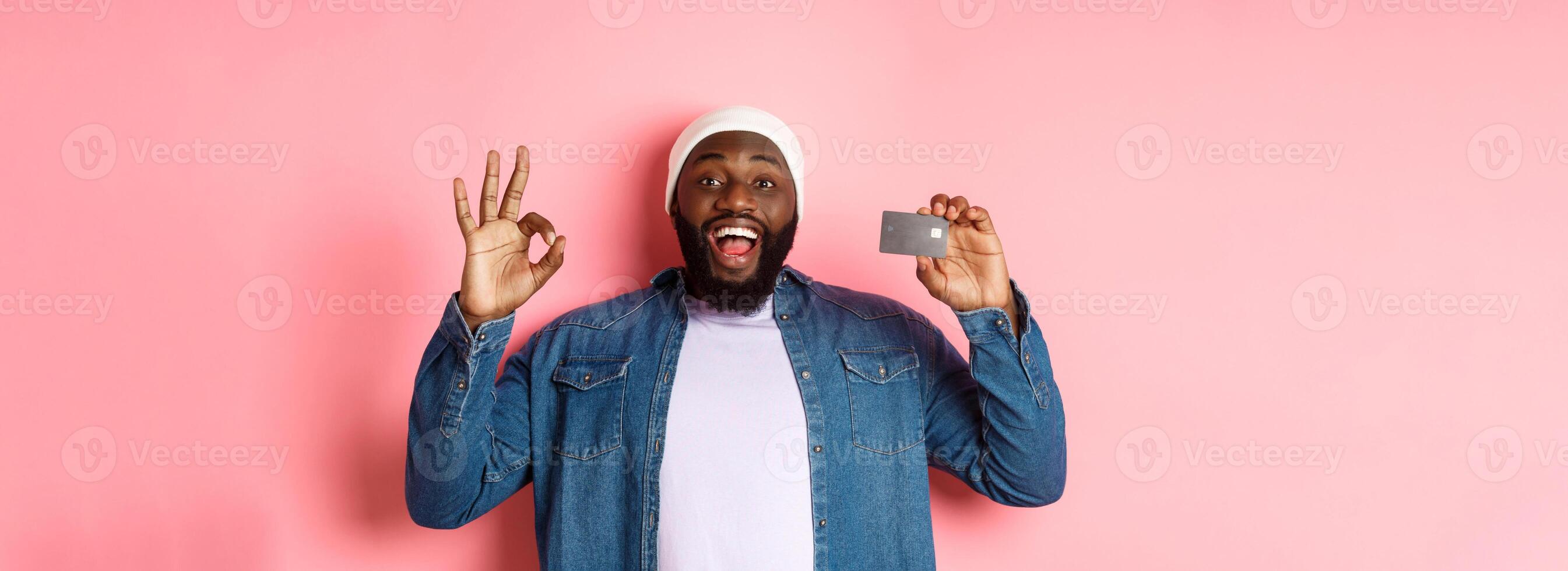 Shopping concept. Handsome african-american man recommending bank, showing credit card and okay sign, smiling satisfied, standing over pink background photo