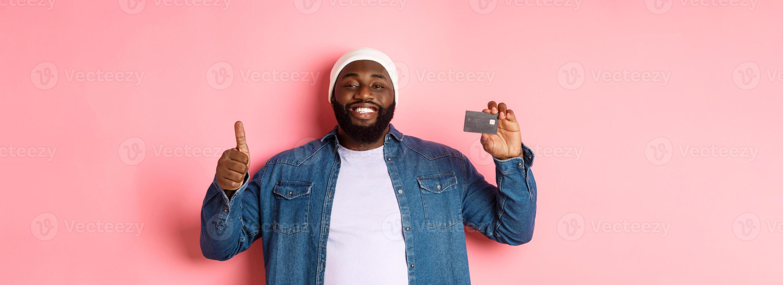 Shopping concept. Handsome african-american man recommending bank, showing credit card and thumbs-up, smiling satisfied, standing over pink background photo