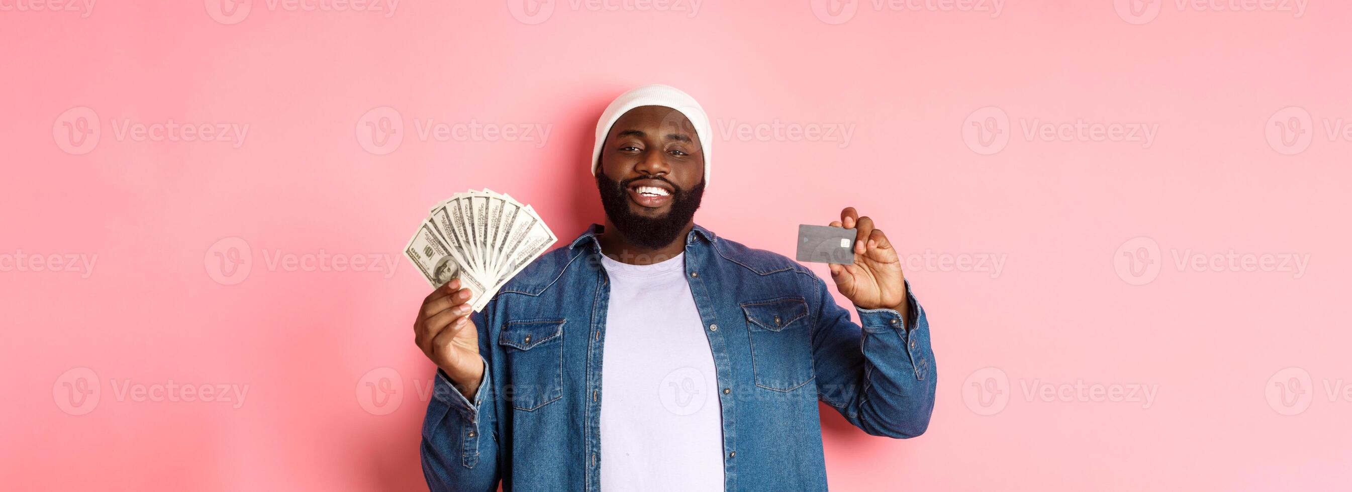 Shopping concept. Handsome young Black guy showing credit card of bank and money, smiling satisfied, standing over pink background photo