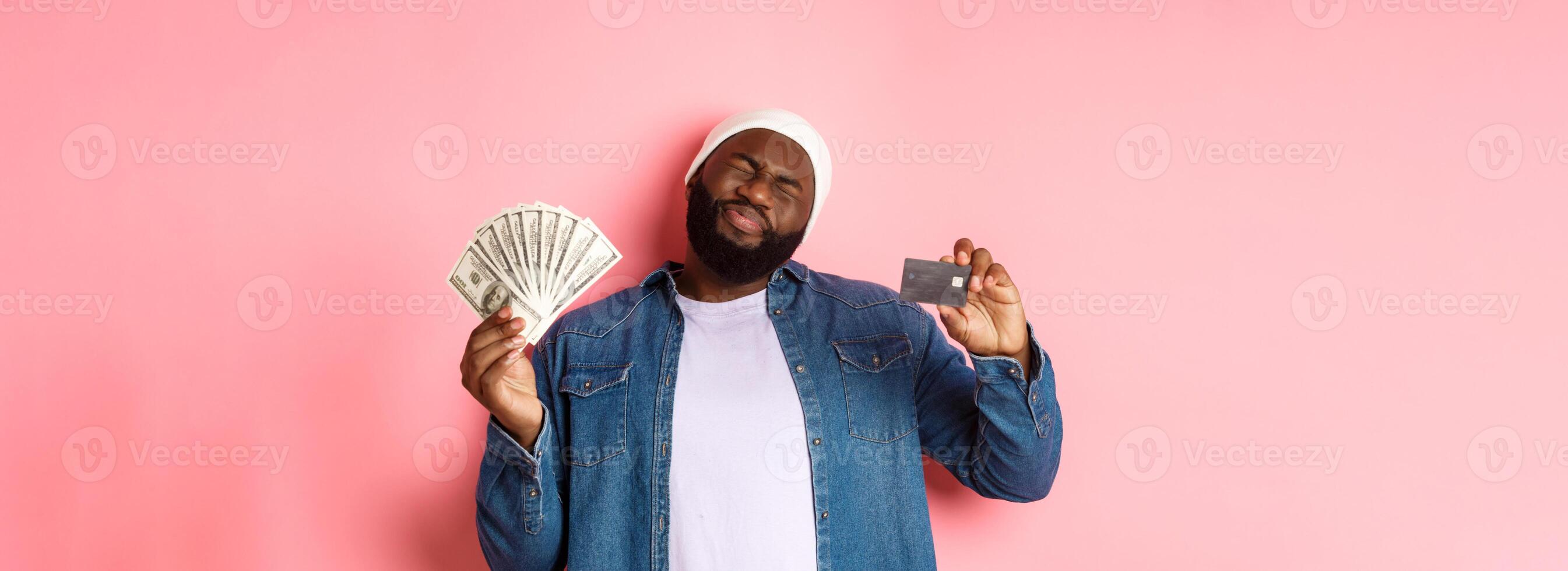 Shopping concept. Sad and whining Black guy showing credit card and dollars, grimacing reluctant, standing over pink background photo