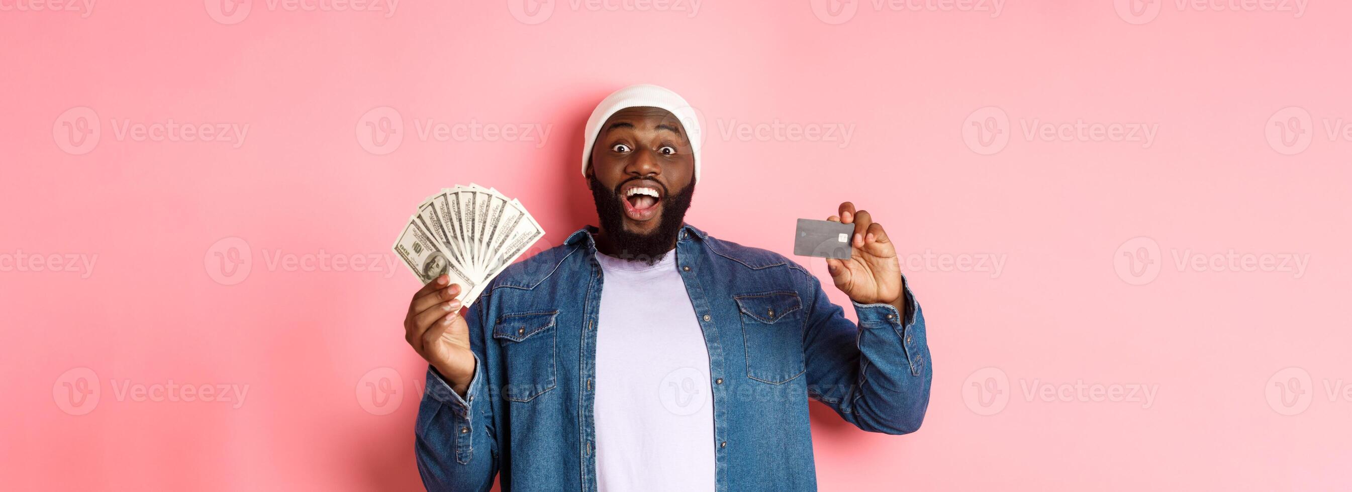 Shopping concept. Excited african-american man showing credit card and dollars, got deposit or money loan, standing over pink background photo