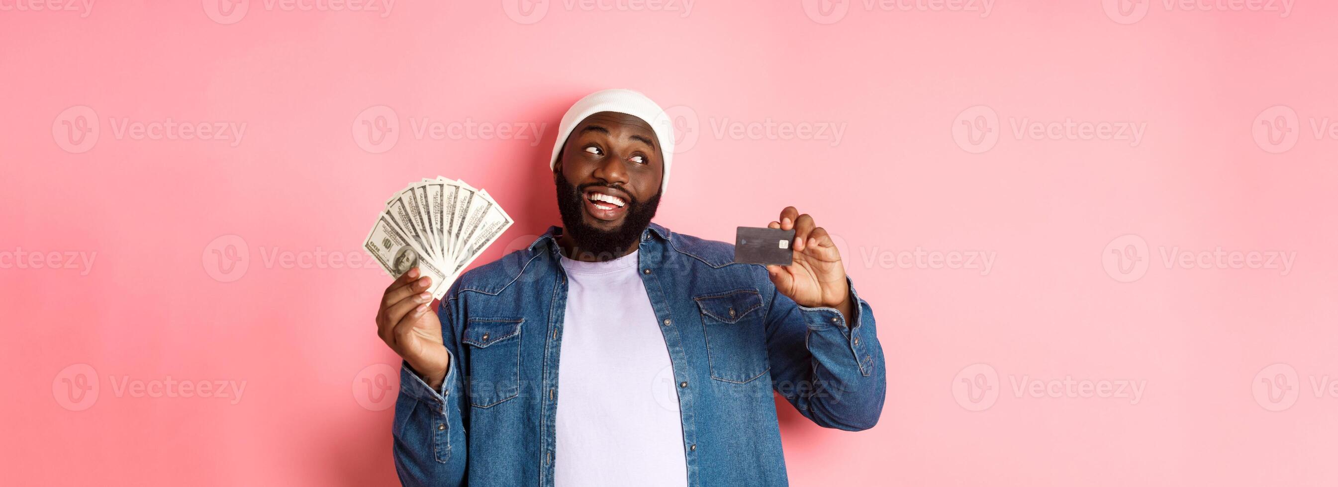 Dreamy african-american man showing credit card and dollars, thinking about shopping and smiling, standing over pink background photo