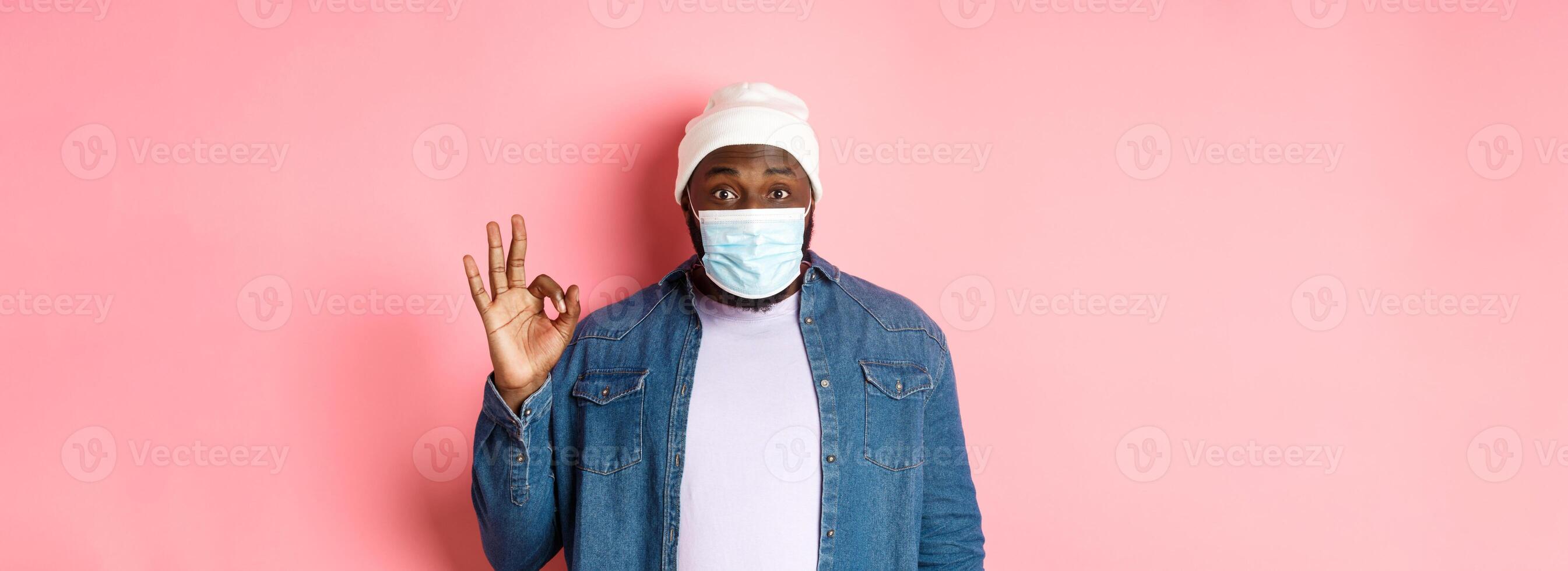 Coronavirus, lifestyle and social distancing concept. Impressed african-american man in face mask, showing okay sign, like and agree, standing over pink background photo
