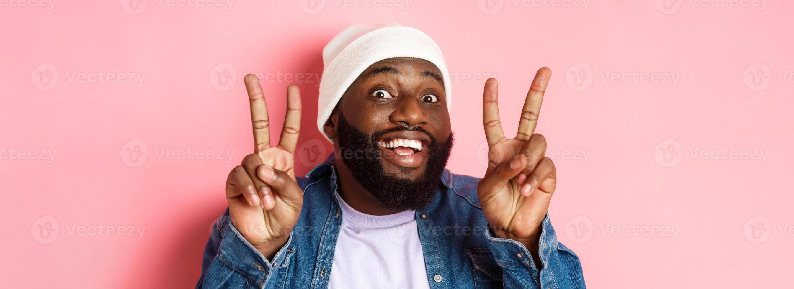 Close-up of cheerful african-american hipster in beanie smiling, showing peace signs, standing happy against pink background photo