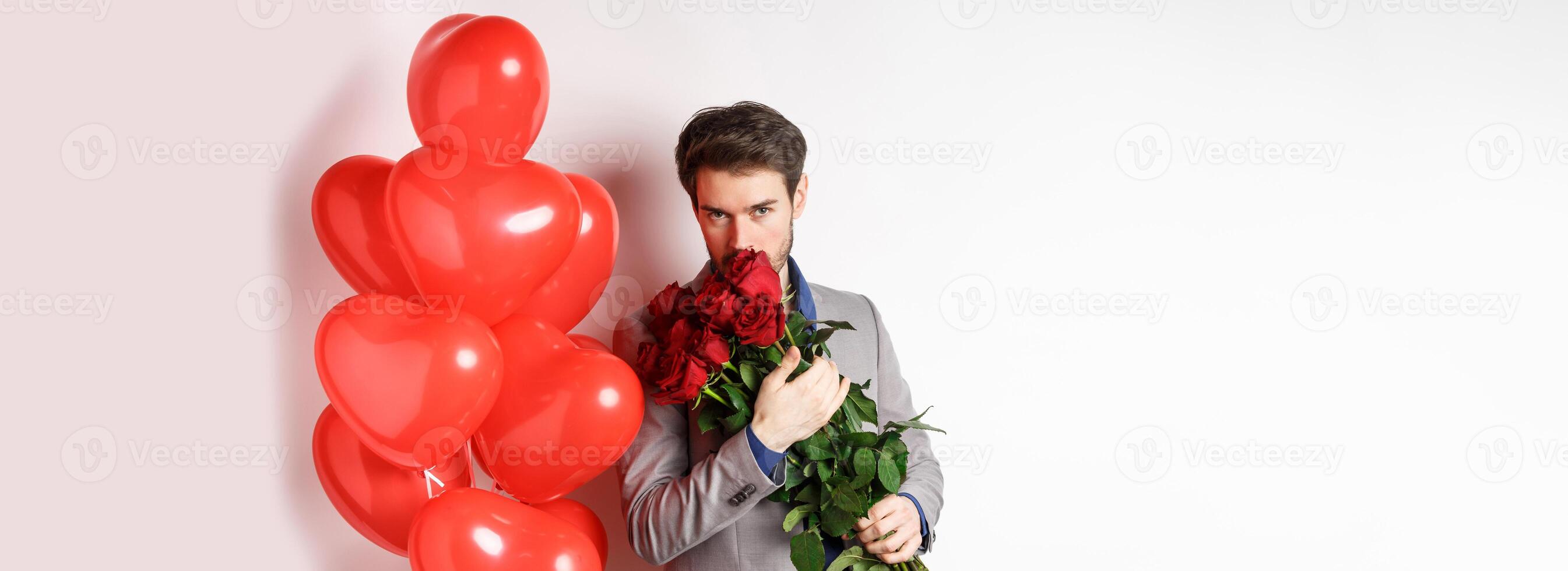 Romantic man smell bouquet of red roses and looking passionate at camera. Boyfriend in suit going on Valentines date with gifts and heart balloons, white background photo